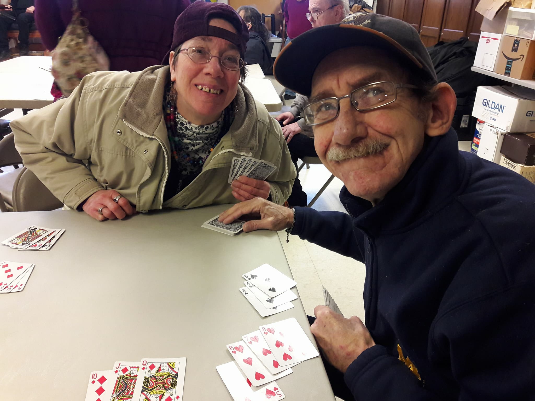 Two people playing a card game smiling at the camera. Both are wearing glasses and hats.