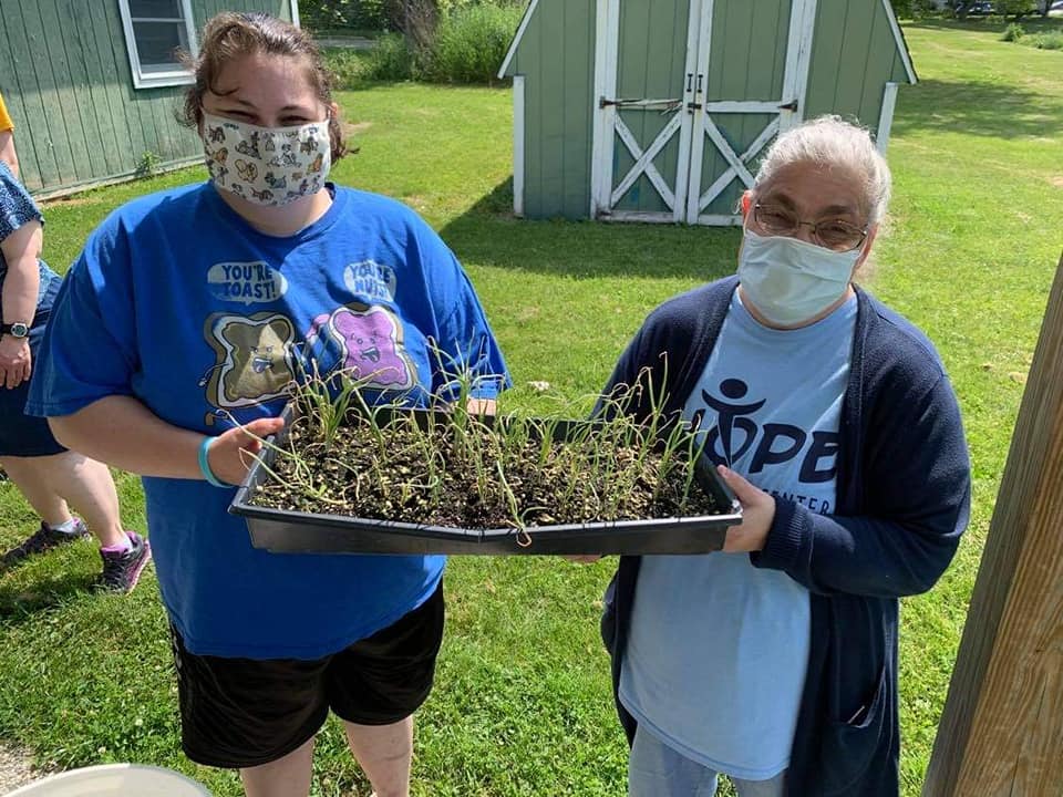 Two people outside wearing masks looking at the camera while holding a tray of sprouting plants.