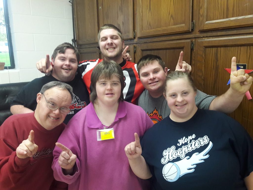 Group of 6 people all standing together smiling and holding up their index fingers to display number 1.