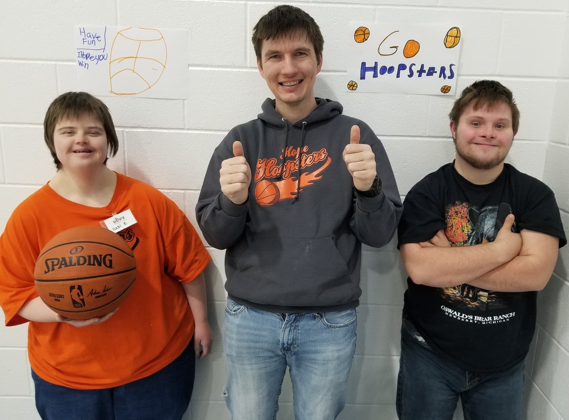 Three people smiling at the camera standing against a white brick wall. One is holding a basketball. One has their thumbs up. One has their arms crossed.