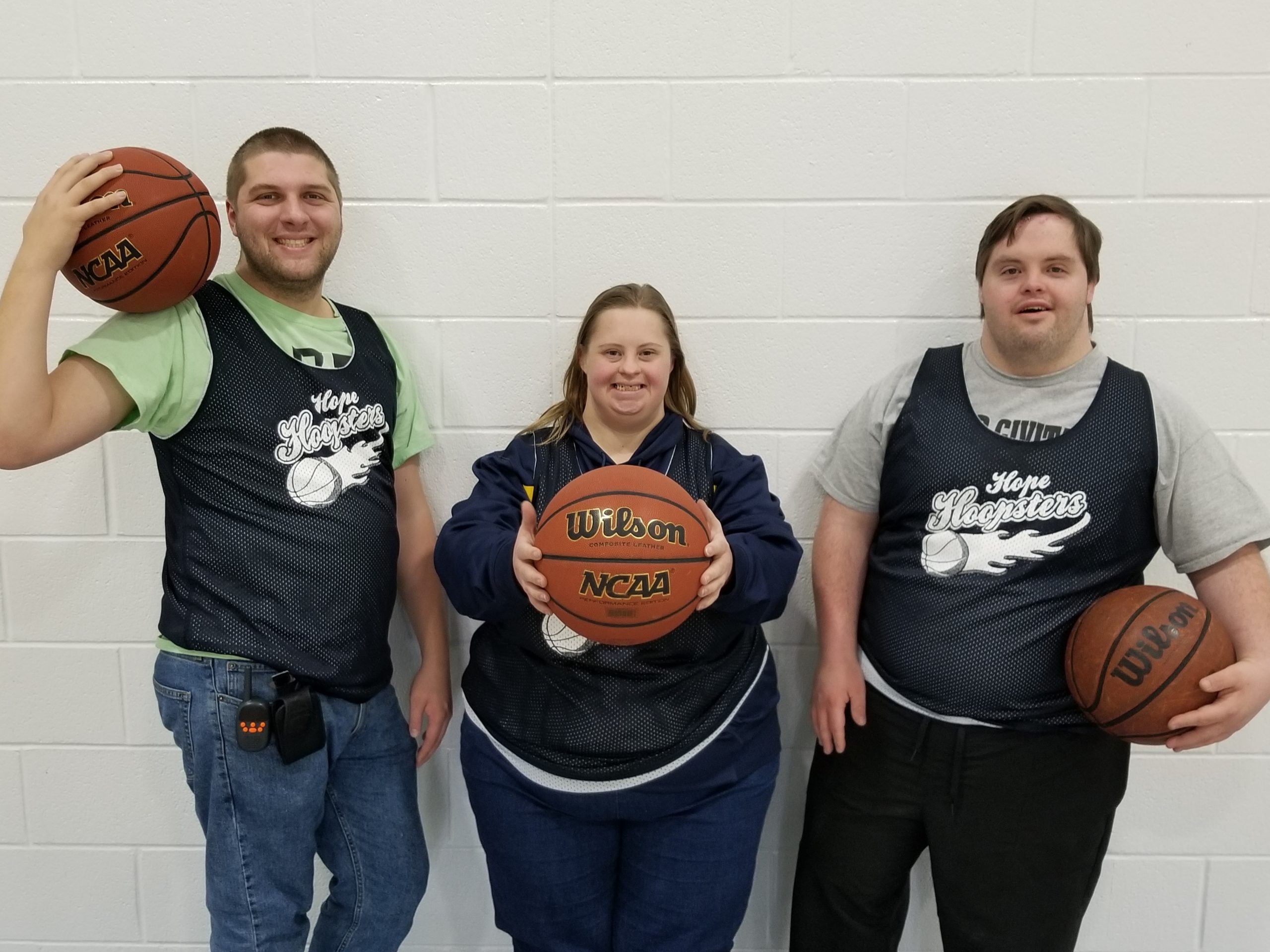 Three people standing together against a white brick wall holding basketballs. They are all wearing blue jerseys that say Hope Hoopsters with a flaming basketball underneath.