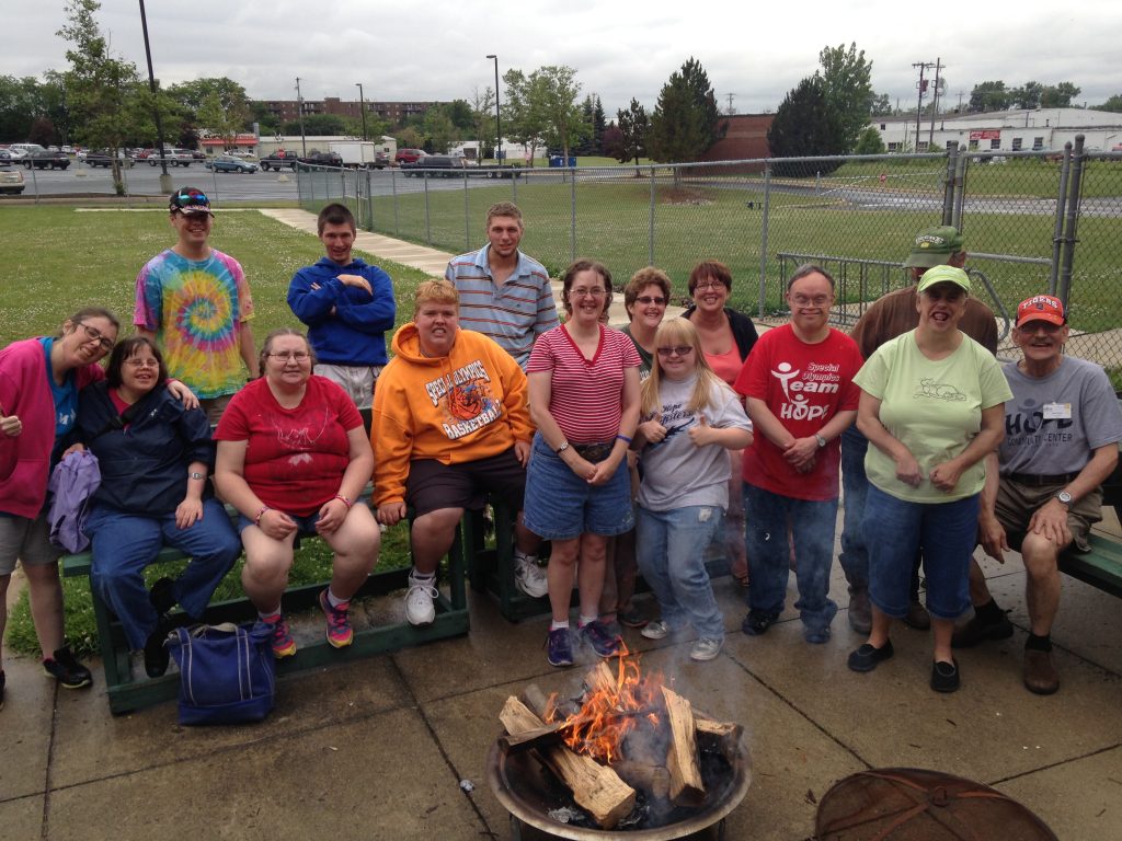 Group of people standing and sitting outside around a fire smiling at the camera.